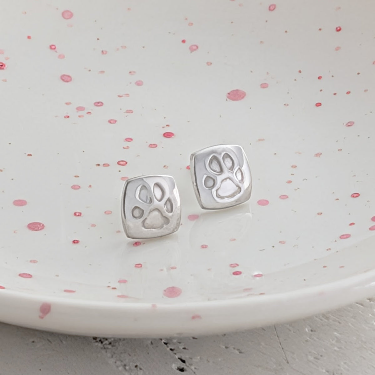Custom pawprint stud earrings crafted from sterling silver, featuring real pet paw prints engraved onto contemporary cushion-shaped studs. Thoughtful pet remembrance jewellery handmade in Shropshire.