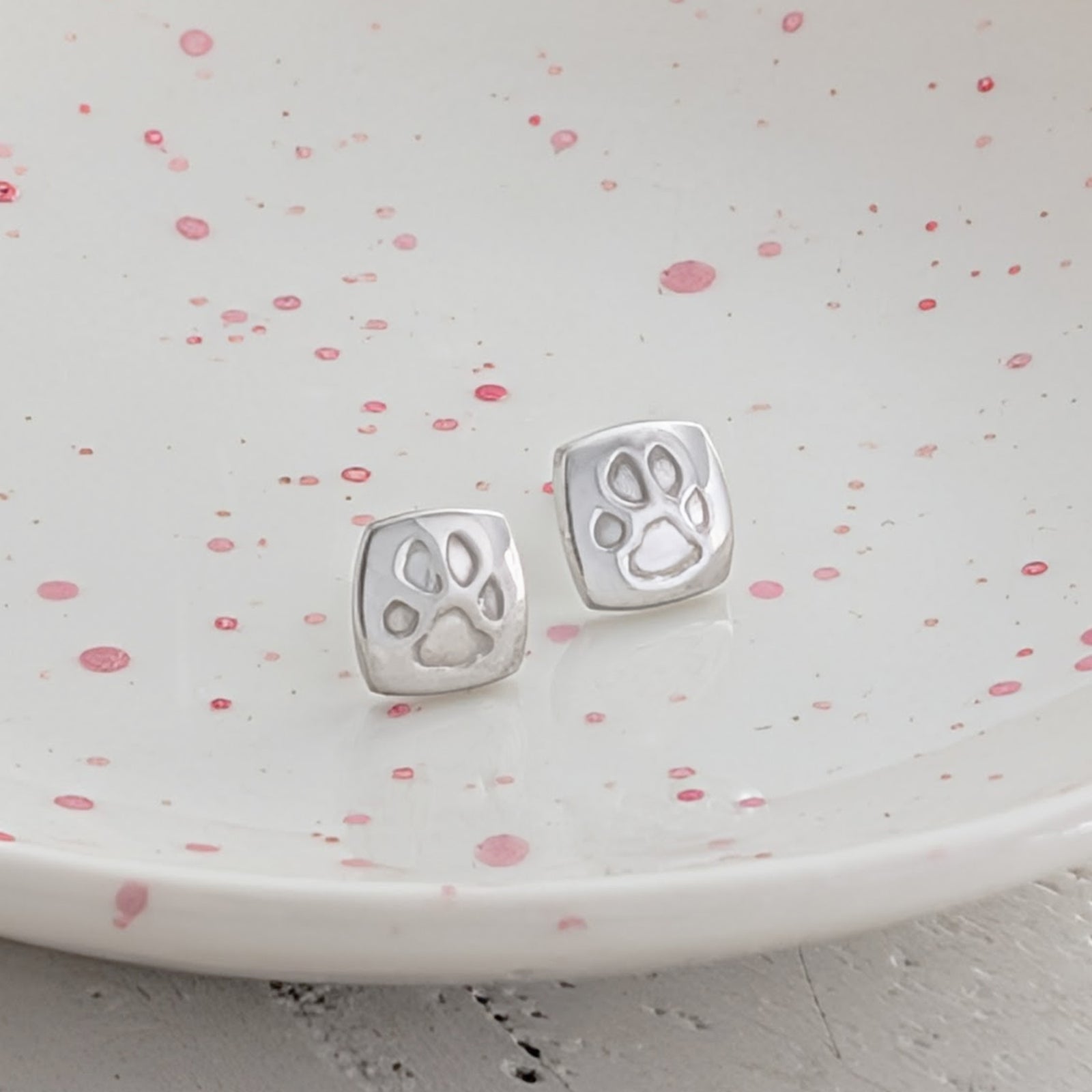 Custom pawprint stud earrings crafted from sterling silver, featuring real pet paw prints engraved onto contemporary cushion-shaped studs. Thoughtful pet remembrance jewellery handmade in Shropshire.